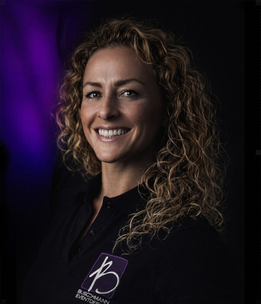 Lächelnde Frau mit lockigem Haar in schwarzem Hemd mit "Buschmann Events"-Logo, lila Licht im Hintergrund.