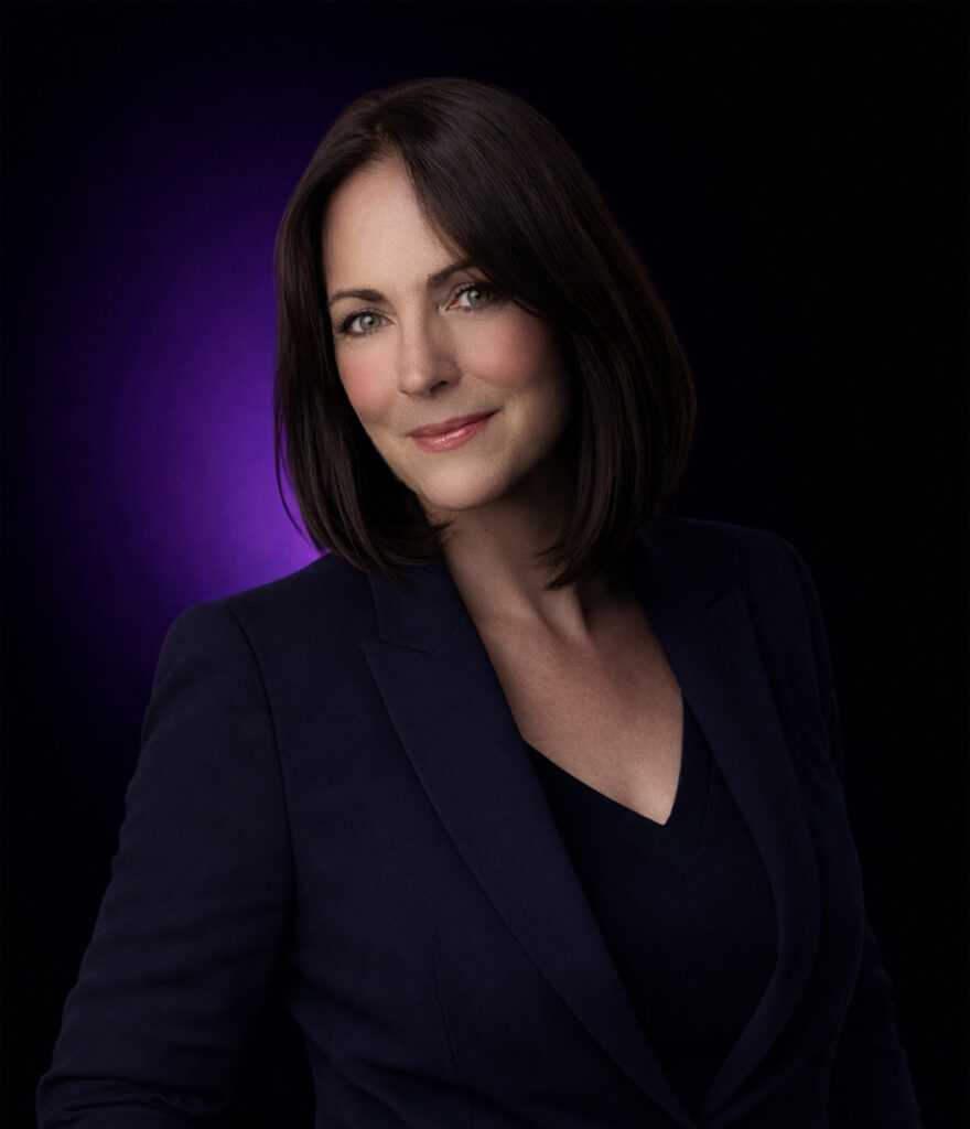 Frau mit schulterlangen braunen Haaren in einem dunklen Anzug, lächelnd, mit lila Hintergrund.