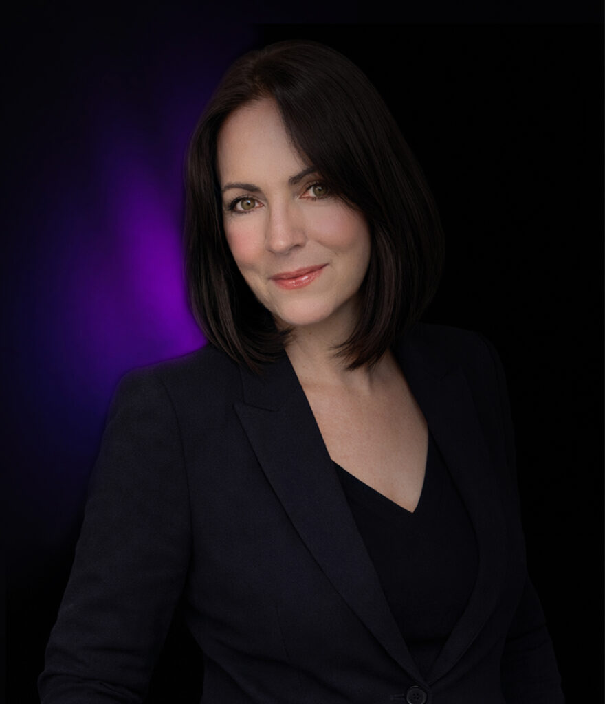 Frau mit schulterlangem dunklem Haar in einem schwarzen Blazer, lächelnd, auf schwarzem Hintergrund mit violettem Licht.