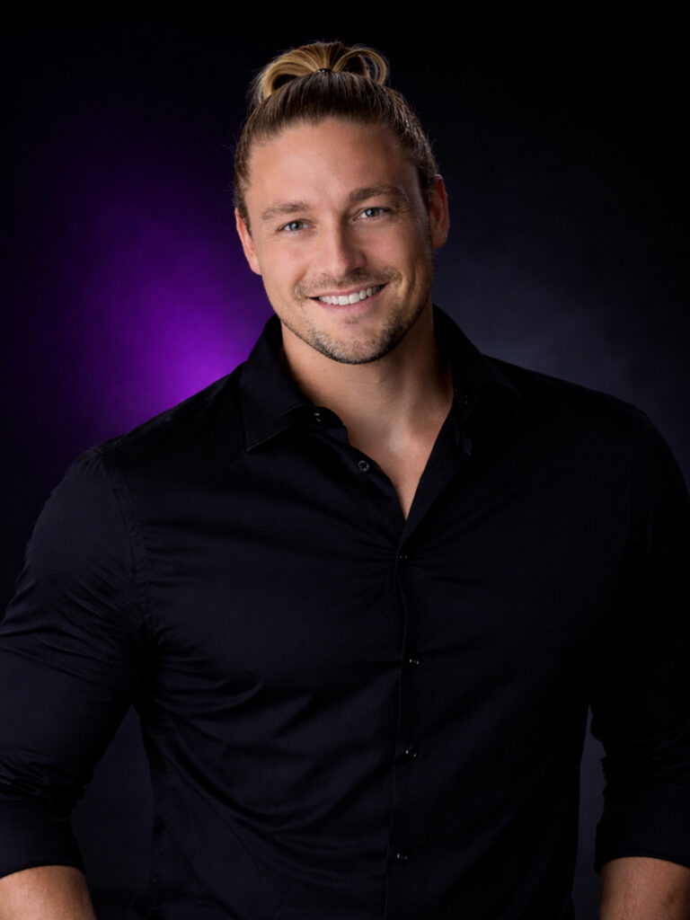 Ein lächelnder Mann mit langen blonden Haaren in einem Dutt, der ein schwarzes Hemd trägt, posiert vor einem dunkelvioletten Hintergrund.